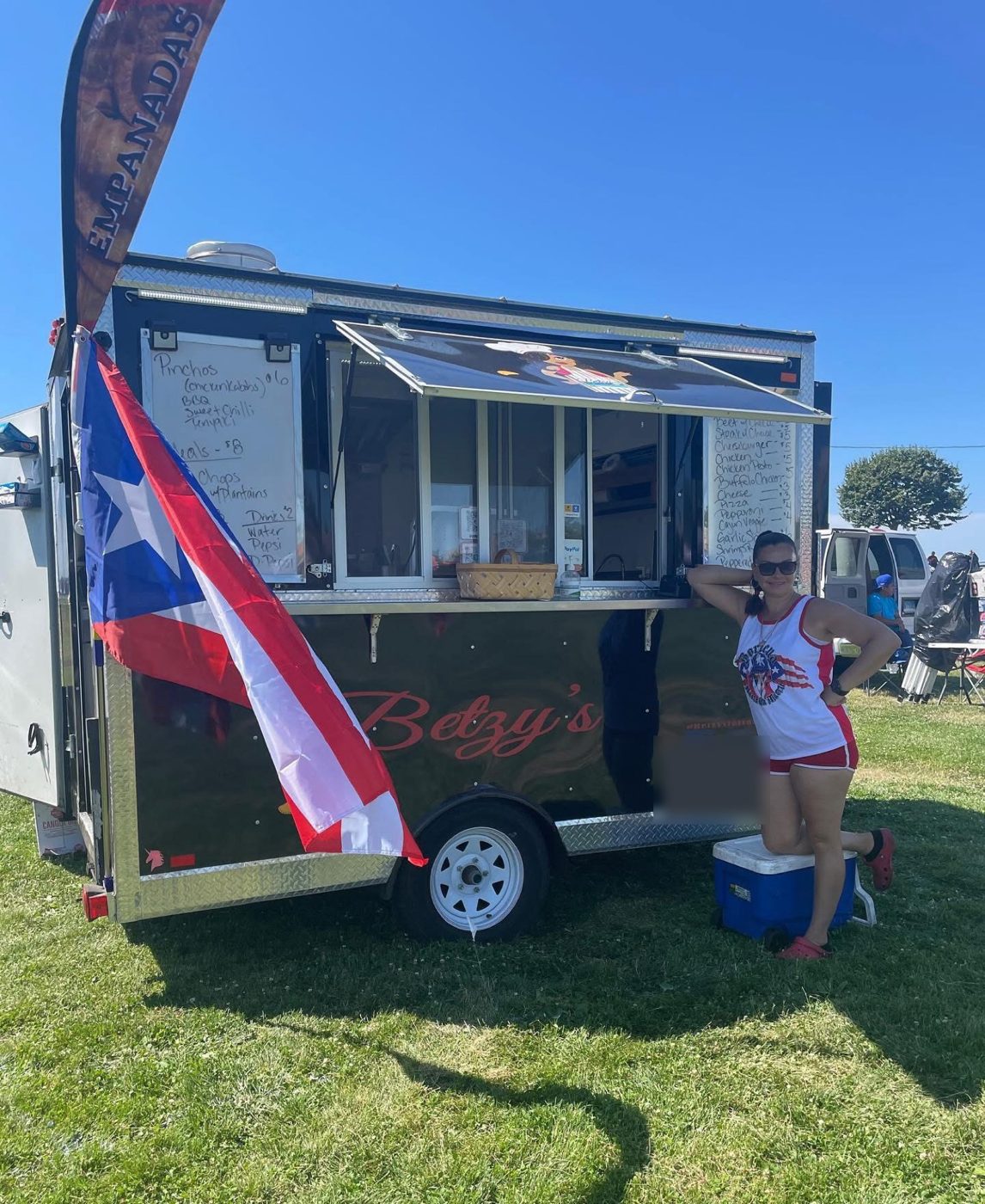 Betzy leaning on her food trailer beside a Puerto Rican flag and an Empanadas feather flag