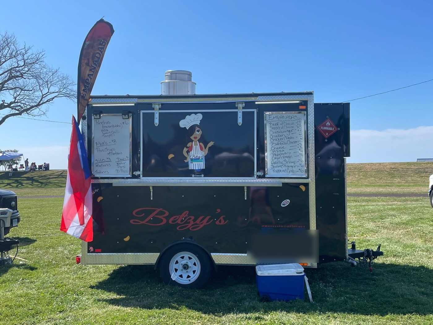 Betzy's black food trailer with a Puerto Rican flag and an Empanadas feather flag, parked on a grassy field